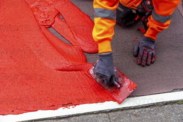 Application of red road surfacing on a cycle lane, inner-city road, so-called cold plastic surfacing, in the colour traffic red, RAL3020, the coloured marking is intended to attract more attention from motorists for cyclists, on cycle paths along roads and at junctions, roundabouts, Essen, North Rhine-Westphalia, Germany