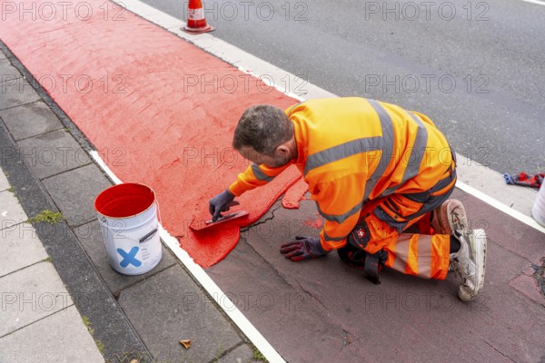 Application of red road surfacing on a cycle lane, inner-city road, so-called cold plastic surfacing, in the colour traffic red, RAL3020, the coloured marking is intended to attract more attention from motorists for cyclists, on cycle paths along roads and at junctions, roundabouts, Essen, North Rhine-Westphalia, Germany