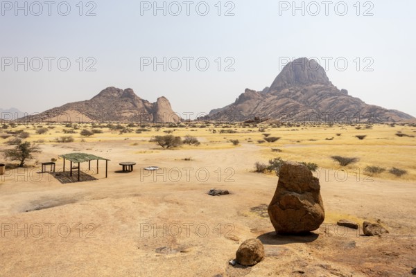 Granite peaks and rugged desert landscape at Spitzkoppe Community Campsite, Namibia. Golden grassland with scattered acacia trees contrasts against dramatic rocky mountains under a clear sky