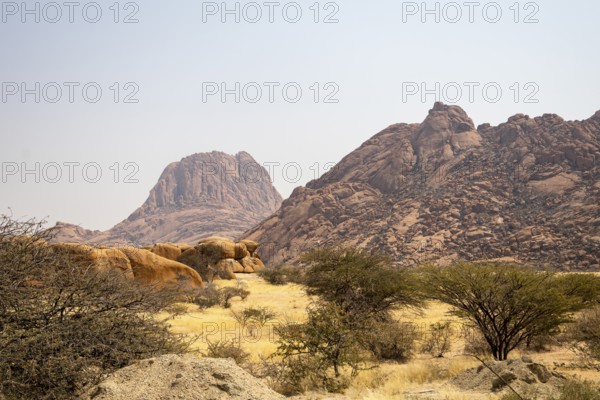 Granite peaks and rugged desert landscape at Spitzkoppe Community Campsite, Namibia. Golden grassland with scattered acacia trees contrasts against dramatic rocky mountains under a clear sky