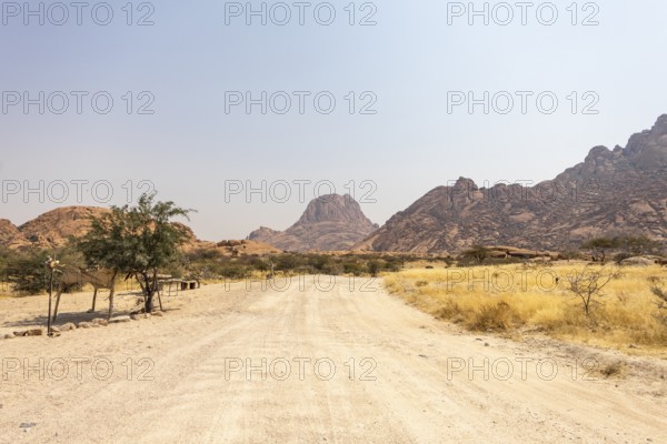 Granite peaks and rugged desert landscape at Spitzkoppe Community Campsite, Namibia. Golden grassland with scattered acacia trees contrasts against dramatic rocky mountains under a clear sky