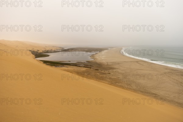 Golden dunes and the Atlantic Ocean near Namib-Naukluft Park, Namibia. A lagoon and shoreline fade into misty, cloudy weather, creating a dramatic desert-sea landscape