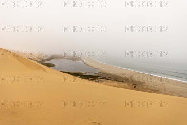 Golden dunes and the Atlantic Ocean near Namib-Naukluft Park, Namibia. A lagoon and shoreline fade into misty, cloudy weather, creating a dramatic desert-sea landscape