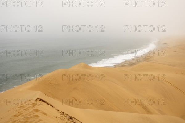 Golden dunes and the Atlantic Ocean near Namib-Naukluft Park, Namibia. A lagoon and shoreline fade into misty, cloudy weather, creating a dramatic desert-sea landscape