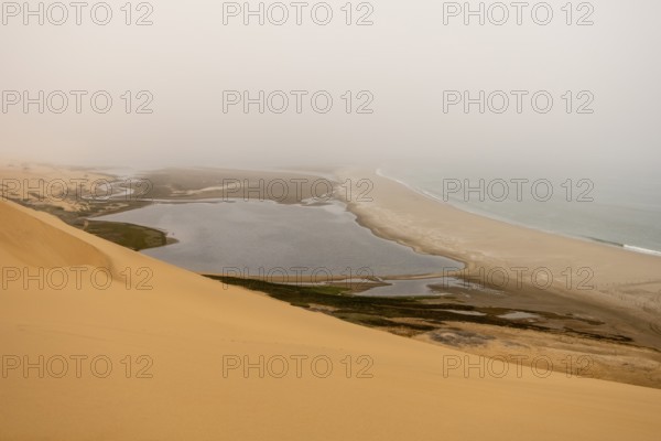 Golden dunes and the Atlantic Ocean near Namib-Naukluft Park, Namibia. A lagoon and shoreline fade into misty, cloudy weather, creating a dramatic desert-sea landscape