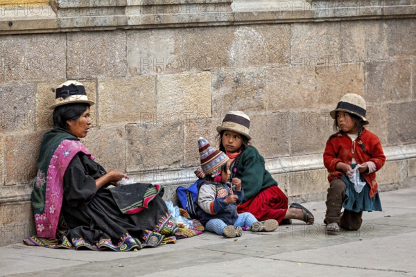 Woman and children in traditional dress sit against a stone wall, lively and colourful, woman and people in the streets of Potosi Bolivia