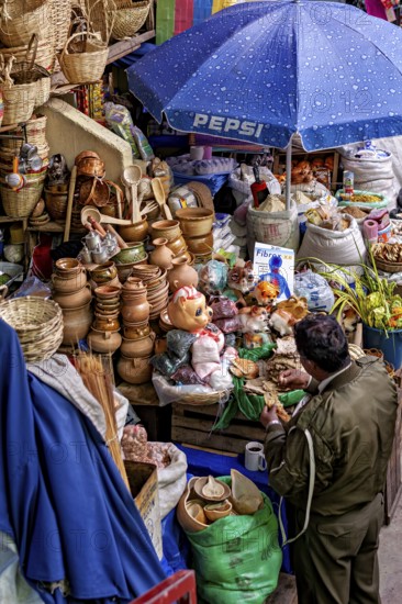 Traders at a lively market with ceramics, dolls and colourful fabrics under a blue umbrella, Extensive range of goods at a market stall in the market halls of Potosi Bolivia