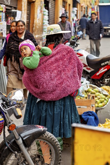 South American street scene at a busy market with traditional clothes and a child on the mother's back, An indigenous woman from Bolivia carries a child through Potosi Bolivia