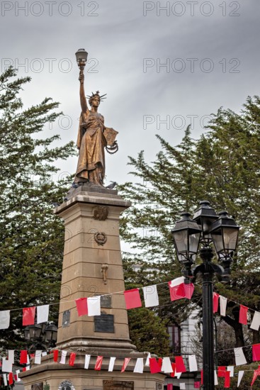 Historical statue with lantern on a high pedestal, surrounded by red and white flags and trees under a cloudy sky, a monument similar to the Statue of Liberty in the city of Potosi in Bolivia