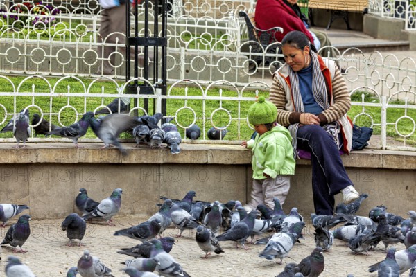 A woman sits with a child in a park, surrounded by many pigeons. There is a calm, everyday atmosphere, A mother with her child in a large group of urban pigeons in Potosi Bolivia