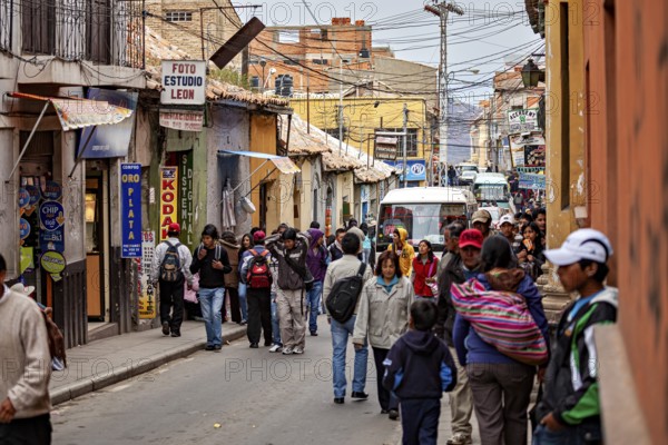 Lots of hustle and bustle in a narrow city street with people walking past. Various shops and colourful signs characterise the lively streetscape, people in the streets of Potosi in Bolivia