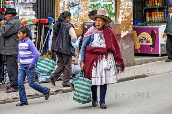 Woman in traditional clothes with bags walking past market stalls on the street, woman and people in the streets of Potosi Bolivia