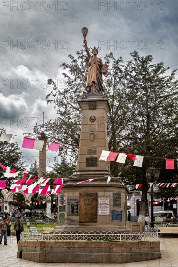 Statue on a stone pedestal in a plaza with red and white flags and trees, under a cloudy sky, Monument of a statue that looks like the Statue of Liberty in Potosi in Bolivia