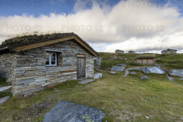 A small stone hut with a grass roof on a rocky meadow, surrounded by other wooden huts and sparse vegetation, Peer Gynt hytta, Rondane National Park, Otta, Innlandet, Norway