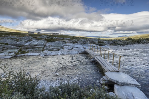 A small wooden bridge crosses a wide river, surrounded by rocks and sparse vegetation in the background, Peer Gynt hytta, Rondane National Park, Otta, Innlandet, Norway