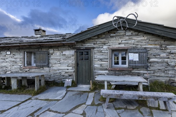 A traditional stone house with a café sign, wooden benches in front, surrounded by a mountainous landscape under a cloudy sky, Peer Gynt hytta, Rondane National Park, Otta, Innlandet, Norway