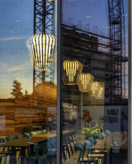 The picture shows the interior of a modern Berlin restaurant, taken in the evening, the scene is characterised by modern lighting fixtures reminiscent of large, glowing light bulbs. Scaffolding can be seen through the glass window, Berlin, Germany