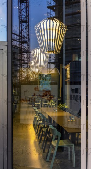 The picture shows the interior of a modern Berlin pub, taken in the evening. The scene is characterised by modern lighting fixtures reminiscent of large, glowing light bulbs. Scaffolding can be seen through the glass window, Berlin, Germany