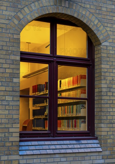 A view through the window of a library, shelves full of books illuminated by a warm, yellow light, Berlin, Germany