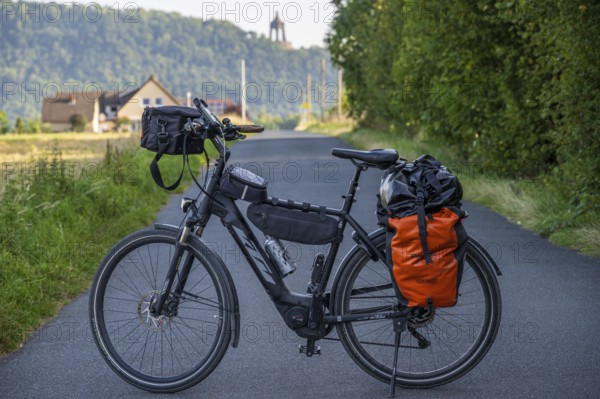 An electric bicycle with an orange bag stands on a rural road, surrounded by green nature and a hill with houses, touring bike in front of the Kaiser Wilhelm monument, Holzhausen, Porta-Westfalica, North Rhine-Westphalia