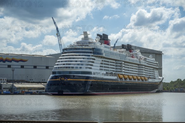 A cruise ship lies in the water in front of a shipyard on a partly cloudy day with a crane in the background, cruise ship in the Meyer shipyard in Papenburg