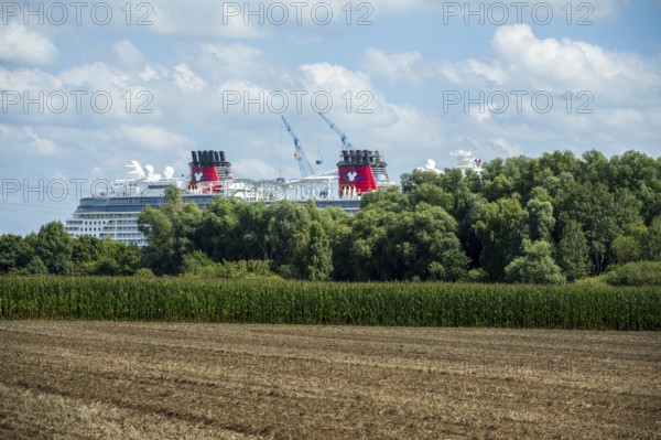 A cruise ship behind trees next to a field under a partly cloudy sky, cruise ship in the shipyard of Papenburg