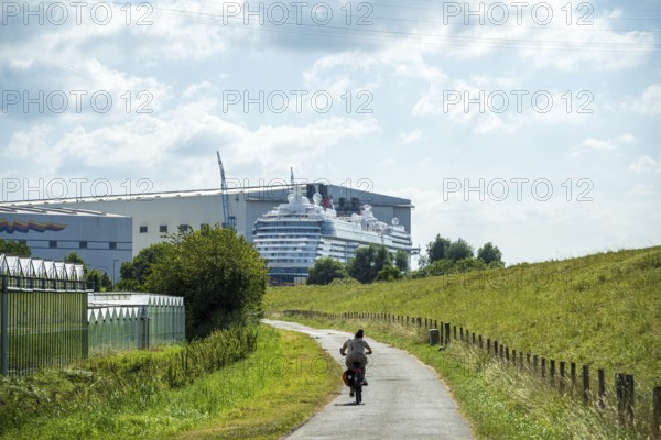 A cyclist on a country road next to a shipyard with a cruise ship in the background, cruise ship at the Meyer shipyard in Papenburg