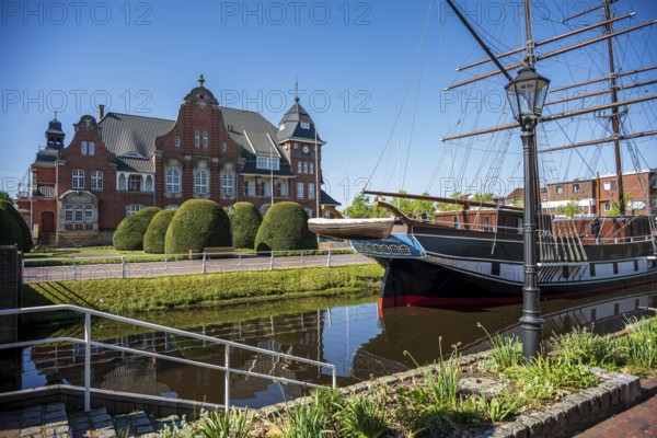 An old ship in front of an impressive brick building, surrounded by manicured nature and water, The sailing ship Frederike von Papenburg in front of the town hall in Papenburg