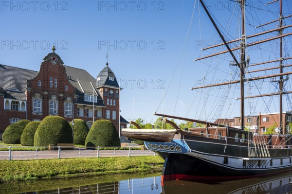 A sailing ship lies in front of a historic building with a blue sky and well-tended bushes on the canal, The sailing ship Frederike von Papenburg in front of the town hall in Papenburg