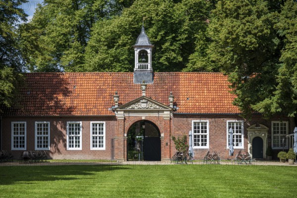 A historic building with a red tiled roof, a small bell tower, surrounded by green trees and a green area in the foreground, The entrance area of Evenburg Castle, Leer, East Frisia