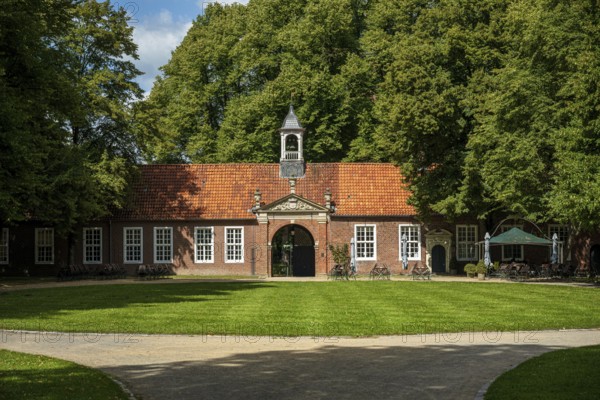 A brick building with a bell tower in front of a grassy area and surrounded by tall trees in the sunny park, The entrance area of Evenburg Castle, Leer, East Frisia