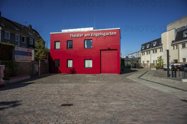 Theatre building in red with cobblestones surrounded by urban elements, high-contrast design, Theater am Engelsgarten in Wuppertal Barmen