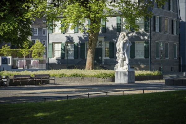 Small square with stone statue in front of an old house with green shutters, illuminated by sunlight, statue The Strong Left by Alfred Hrdlicka in front of the Friedrich Engels House in Wuppertal Barmen