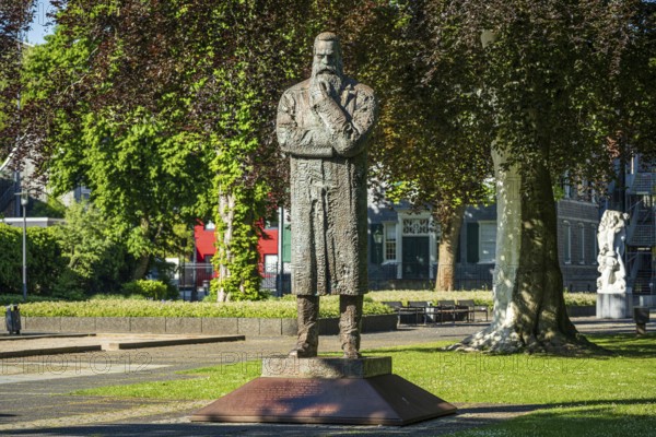 Stone statue of a man with crossed arms in an urban park with surrounding buildings, statue of Friedrich Engels by sculptor Zeng Chenggang in the Engelsgarten Wuppertal Barmen
