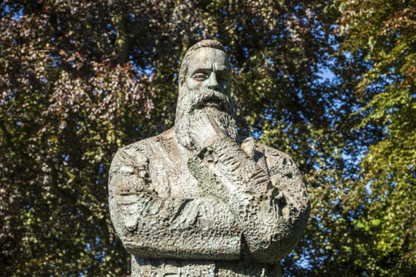 Close-up of a stone statue of a man with a beard, surrounded by trees and a clear sky, statue of Friedrich Engels by sculptor Zeng Chenggang in the Engelsgarten Wuppertal Barmen
