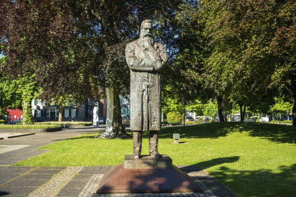 Stone statue of a man with crossed arms in a sunny park with many trees, statue of Friedrich Engels by sculptor Zeng Chenggang in the Engelsgarten Wuppertal Barmen