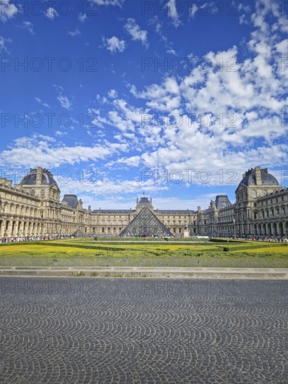A grand panoramic view of the Louvre Palace with its iconic glass pyramid in the center under a magnificent blue sky with fluffy white clouds in Paris, France