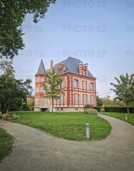 The Castel Rose in L'Isle-Adam, France. Historic mansion characterized by its pink walls and distinctive red brick trim. The building is surrounded by a lush green lawn and winding paths