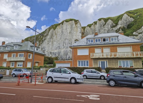 Row of houses with distinctive orange brickwork sits at the base of dramatic white chalk cliffs in Dieppe, France. Parked cars along the street with a red-toned bike lane in the foreground