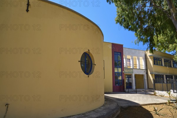 Gymnasium, Yellow modern building with round shapes, next to a tree under a blue sky, Historicism, Art Deco, Rationalism, Portolago, Lakki, Leros, Dodecanese, Greek Islands, Greece