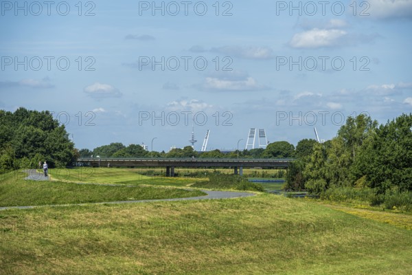 A bridge leads over green areas with trees and a path under a blue sky with few clouds. Stadium towers are visible, Weser cycle path, Weser, floodlight masts Weserstadion, Bremen