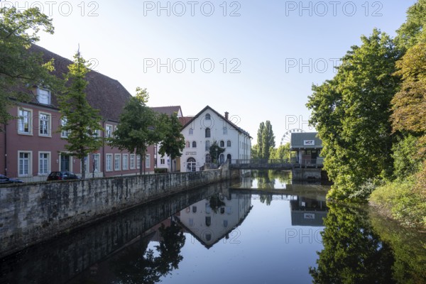 River reflects buildings and trees under a clear sky with a Ferris wheel in the background, Warendorf, Münsterland, North Rhine-Westphalia