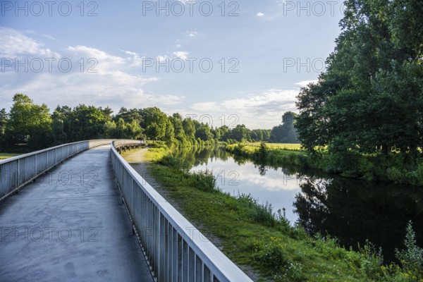 Path along a calm river, lush vegetation and cloudy sky are reflected in the water, Emssee near Warendorf, Münsterland