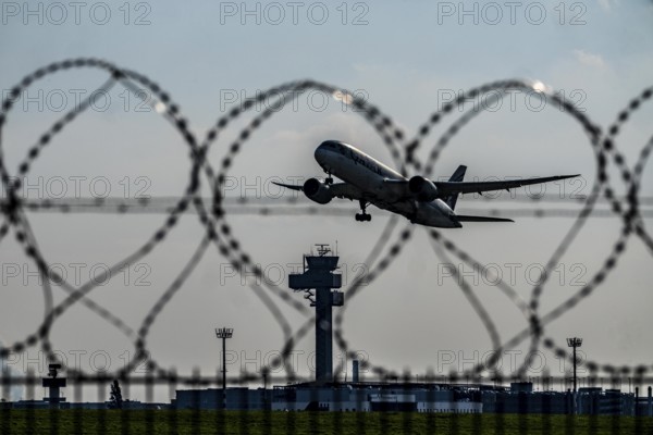 Symbolic image security at the airport, outer fence at Düsseldorf International Airport, steel wire fence, with S-wire rolls, NATO wire, on the top of the fence, aircraft taking off, air traffic control tower, DFS, North Rhine-Westphalia, Germany