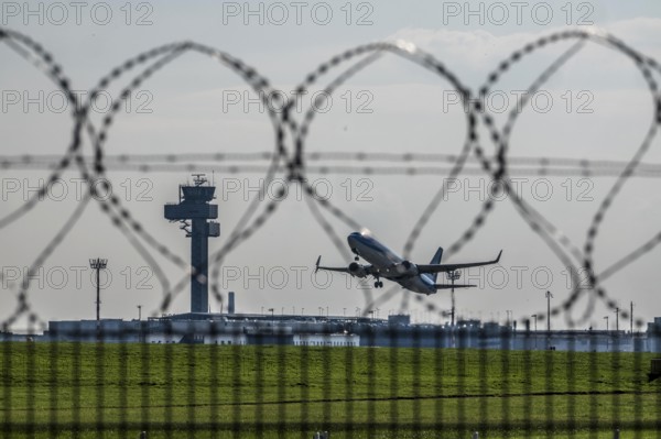Symbolic image security at the airport, outer fence at Düsseldorf International Airport, steel wire fence, with S-wire rolls, NATO wire, on the top of the fence, aircraft taking off, air traffic control tower, DFS, North Rhine-Westphalia, Germany