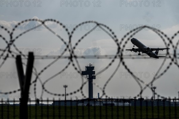 Symbolic image security at the airport, outer fence at Düsseldorf International Airport, steel wire fence, with S-wire rolls, NATO wire, on the top of the fence, aircraft taking off, air traffic control tower, DFS, North Rhine-Westphalia, Germany