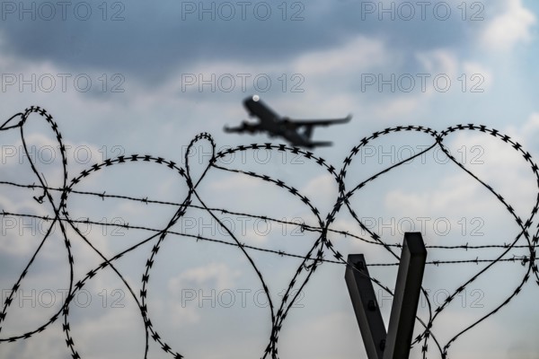 Symbolic image security at the airport, outer fence at Düsseldorf International Airport, steel wire fence, with S-wire rolls, NATO wire, on the top of the fence, aeroplane taking off, North Rhine-Westphalia, Germany