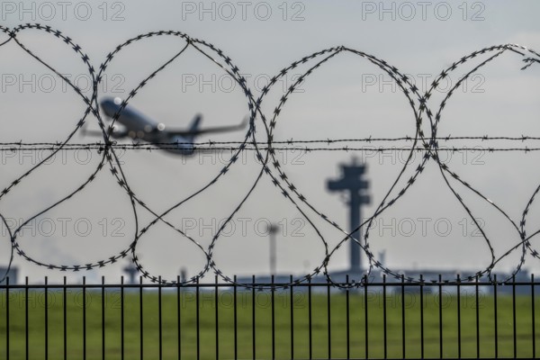 Symbolic image security at the airport, outer fence at Düsseldorf International Airport, steel wire fence, with S-wire rolls, NATO wire, on the top of the fence, aircraft taking off, air traffic control tower, DFS, North Rhine-Westphalia, Germany