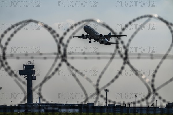 Symbolic image security at the airport, outer fence at Düsseldorf International Airport, steel wire fence, with S-wire rolls, NATO wire, on the top of the fence, aircraft taking off, air traffic control tower, DFS, North Rhine-Westphalia, Germany