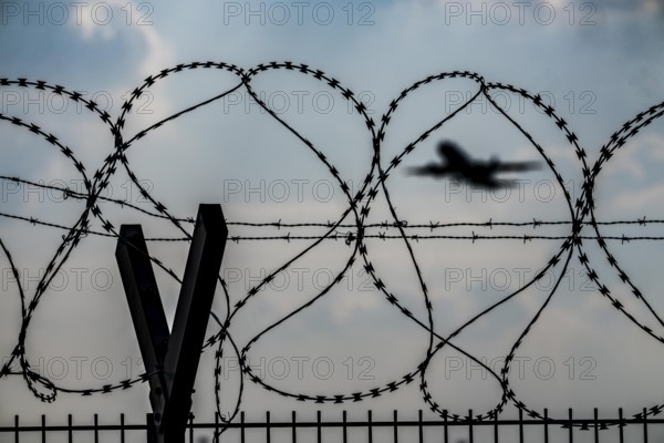 Symbolic image security at the airport, outer fence at Düsseldorf International Airport, steel wire fence, with S-wire rolls, NATO wire, on the top of the fence, aeroplane taking off, North Rhine-Westphalia, Germany